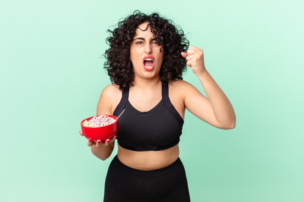 Pretty arab woman shouting aggressively with an angry expression wearing fitness clothes and holding a corn flakes bowl