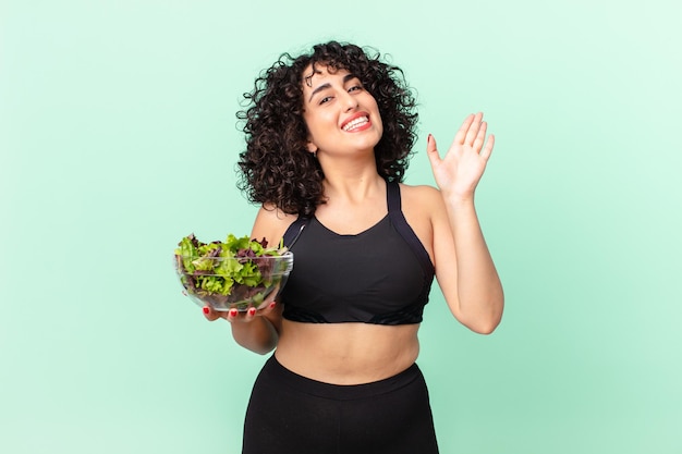 Pretty arab woman smiling happily, waving hand, welcoming and greeting you and holding a salad. diet concept