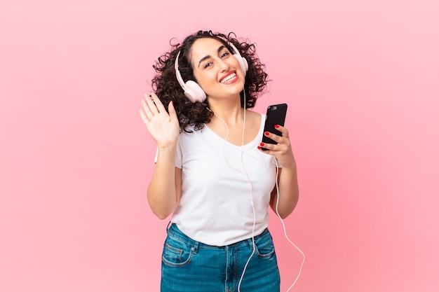 Pretty arab woman smiling happily, waving hand, welcoming and greeting you with headphones and a smartphone