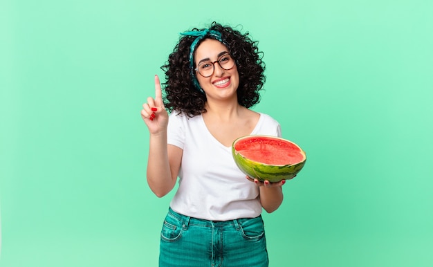 Pretty arab woman smiling and looking friendly, showing number one and holding a watermelon. summer concept