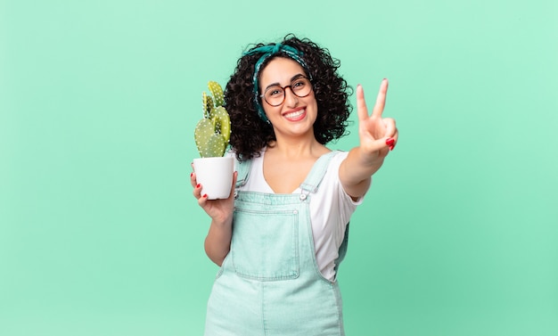 Pretty arab woman smiling and looking happy, gesturing victory or peace and holding a potted cactus