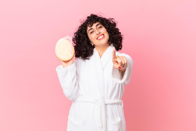 Pretty arab woman smiling proudly and confidently making number one wearing bathrobe and holding a sponge