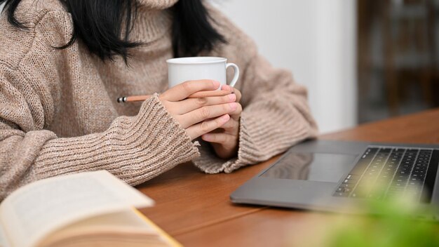 A pretty Asian female in comfy sweater holding a coffee mug working on laptop