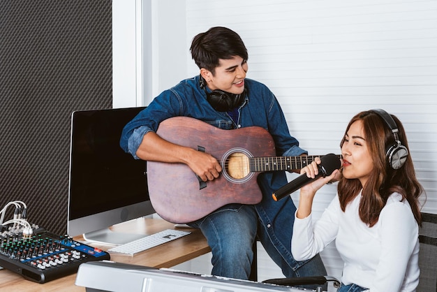 Pretty Asian female singer recording songs by using a studio microphone with male playing guitar Performance and show in the music business Duet session