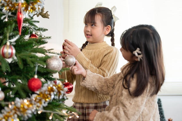 Pretty Asian girl is enjoying decorating a Christmas tree with her younger sister in the living room