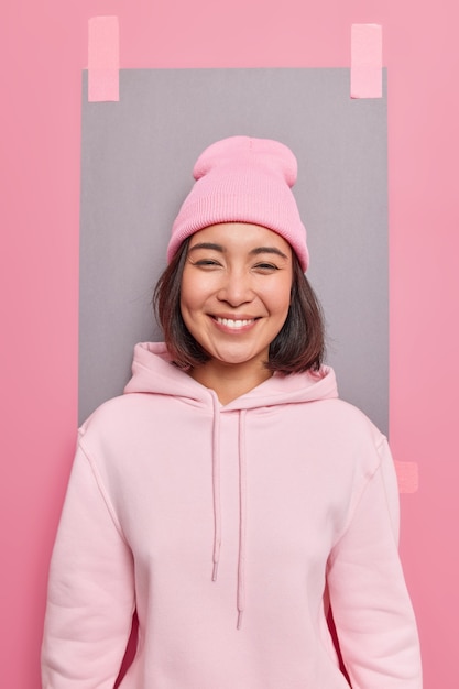 Pretty Asian girl with gentle smile has positive attitude wears sweatshirt and hat glad to hear good news poses against pink wall plastered grey sheet of paper to write your information