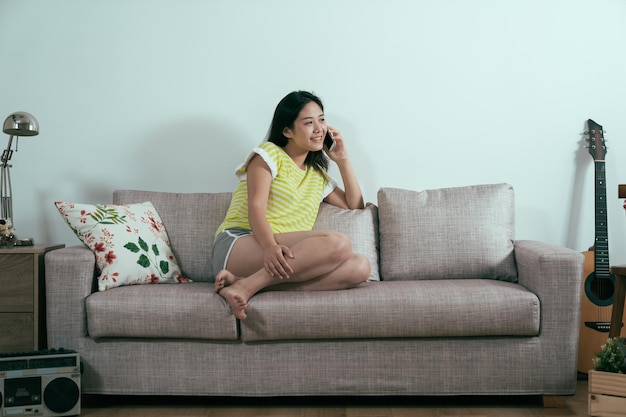 pretty asian korean woman talking on smart phone sitting relaxed on comfort sofa smiling happy laughing on couch relaxing with legs up at home living room. joyful lady chatting gossip on cellphone