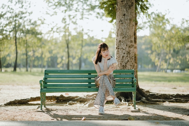 그녀의 다리와 함께 앉아 흰 옷을 입고 꽤 아시아 아가씨는 편안하고 그녀의 전화에서 음악을 듣고 벤치에 넘어