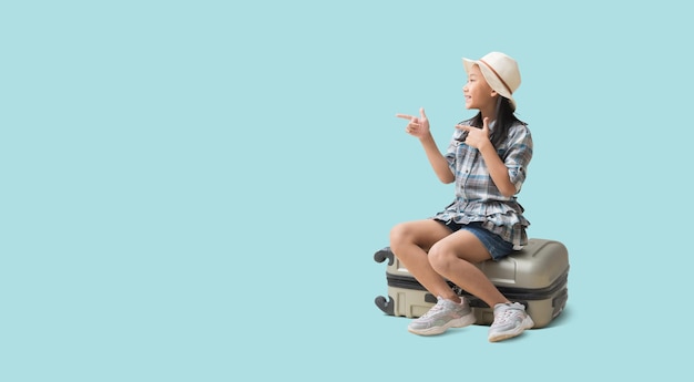 Красивая азиатская маленькая девочка сидит на чемодане с указательным пальцем на пустое пространство Приключенческий отпуск, путешествие, путешествие, мечта, концепция, изолированная на синем фоне с обтравочными дорожками для дизайнерских работ