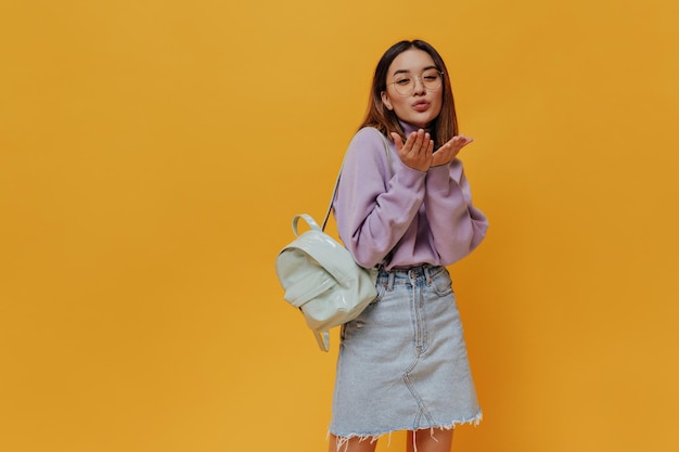 Pretty Asian woman in eyeglasses denim skirt and purple sweater blows kiss and poses with mint backpack on orange background