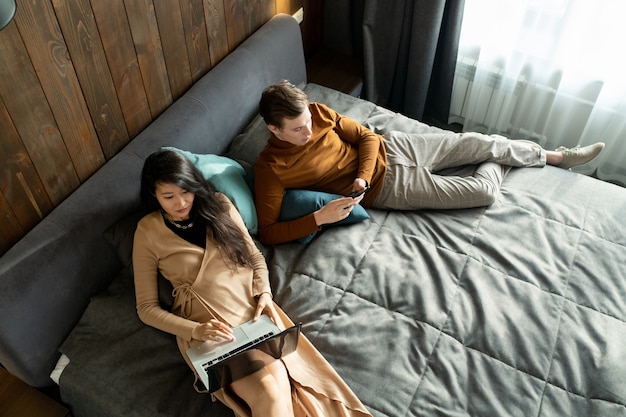 写真 彼女の夫またはボーイフレンドがスマートフォンでスクロールしながら、ラップトップを使用してエレガントなベージュのドレスを着たきれいなアジアの女性、両方ともホテルの部屋のベッドに横たわっています