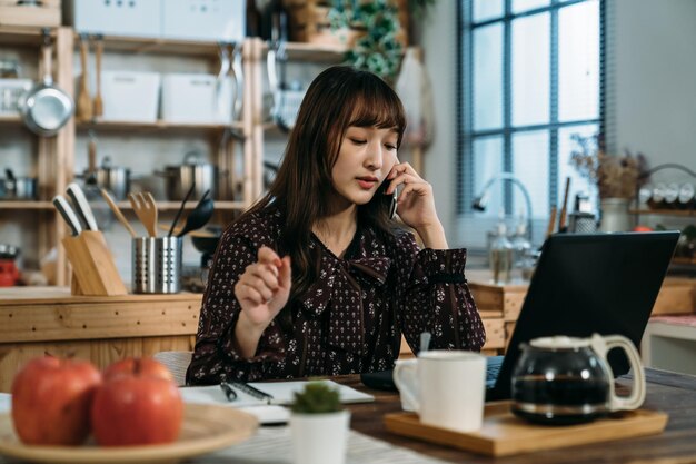 Pretty Asian woman is using mobile phone to discuss the new itinerary with friends for the upcoming holiday next week in the kitchen with hot coffee. work at home concept