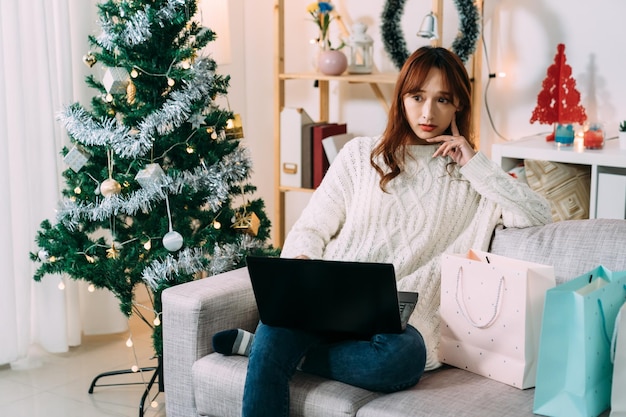 pretty asian woman looking into distance is thinking about what to buy while shopping online for Christmas gift on the computer in the living room at home.