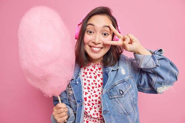 Pretty Asian woman makes v sign over eye has fun enjoys listening music wears headphones holds sweet candy floss dressed in fashionable outfit enjoys free time being in good mood poses indoor