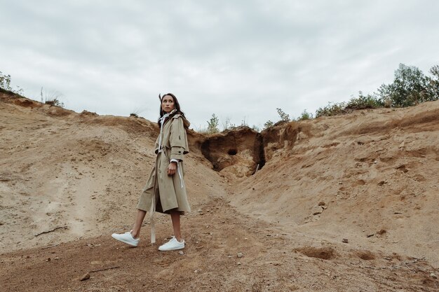 Pretty Asian woman posing full-height in the sand quarry in her beige cloak.