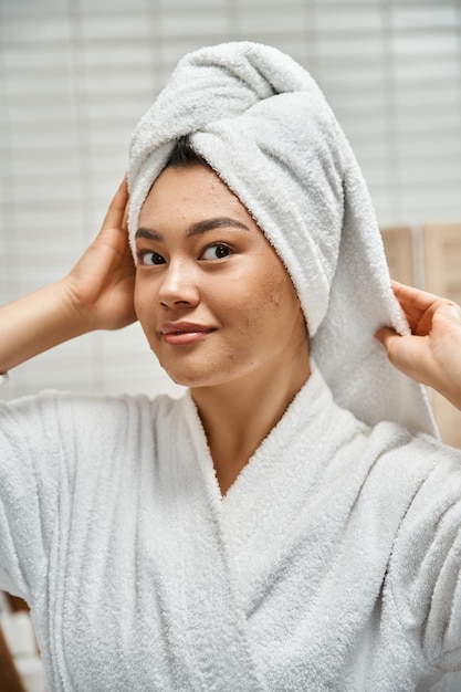 pretty asian woman in robe with white towel on head looking at camera in bathroom skin issues