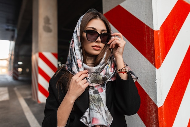 Pretty attractive fashion model young woman in fashionable sunglasses in stylish silk scarf on head in coat posing outdoors near the modern red-white column. Business girl professional in the city.