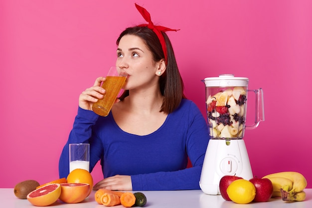 Donna abbastanza attraente con la fascia rossa sulla sua testa che beve frullato di frutta arancione, alzando lo sguardo, tenendo il bicchiere