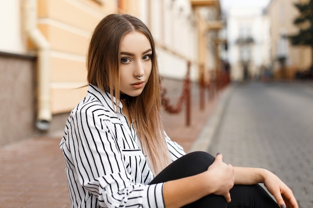 Giovane donna abbastanza attraente in una camicetta a righe bianco e nero vintage in jeans alla moda con eleganti collane di velluto si siede in città in una calda giornata primaverile.