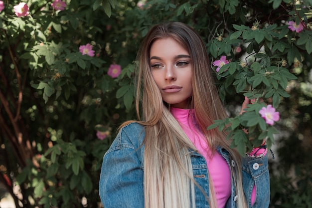 Довольно привлекательная молодая женщина с роскошными длинными волосами в винтажной джинсовой куртке в стильном розовом топе возле зеленого цветущего куста в саду. красивая гламурная девушка расслабляется на открытом воздухе в летний день