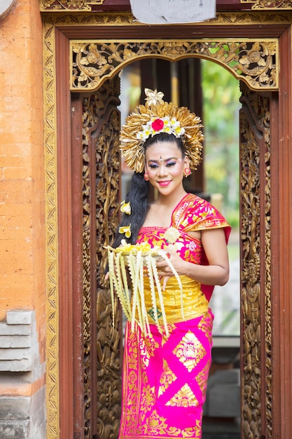 Pretty balinese woman with traditional clothes holding a bowl of offerings and smiling on the temple door