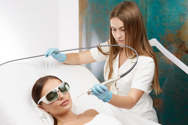 Pretty beautician doing facial procedures with laser apparatus for beautiful female customer