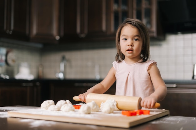 Pretty beautiful little girl is rolling the dough out with wooden rolling pin at the table in kitchen with modern interior, looking at the camera. Face of baby in flour.