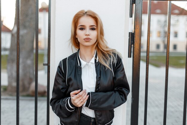 Pretty beautiful young blond woman in a black spring stylish jacket in black jeans in white shirt stands near the vintage iron gate at sunset. Fashionable girl. American fashion