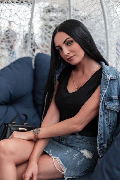 Photo pretty beautiful young brunette woman in a fashionable denim jacket in a stylish black t-shirt in a trendy blue skirt sits on a vintage armchair in an outdoor cafe. cute girl is resting on the street.