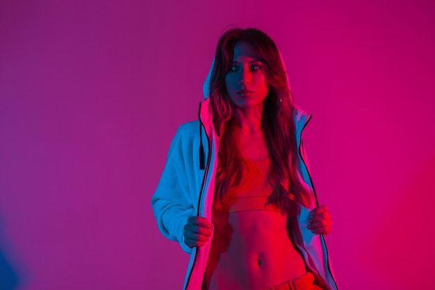 Pretty beautiful young woman in a stylish top in a hoodie with a hood posing in the studio with a brightly neon red light. Fashionable attractive girl fashion model indoors with multi-colored glitter.