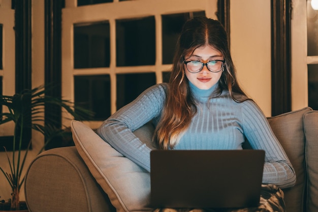 Pretty and beautiful young woman using laptop and surfing on the net at late night on the sofa at home Attractive caucasian teenager girl with computer online Lady wearing glasses working