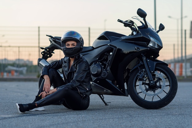 Pretty biker woman with beautiful eyes in black leather jacket and protective helmet sits near stylish sports motorcycle at urban parking. Traveling and active lifestyle concept.