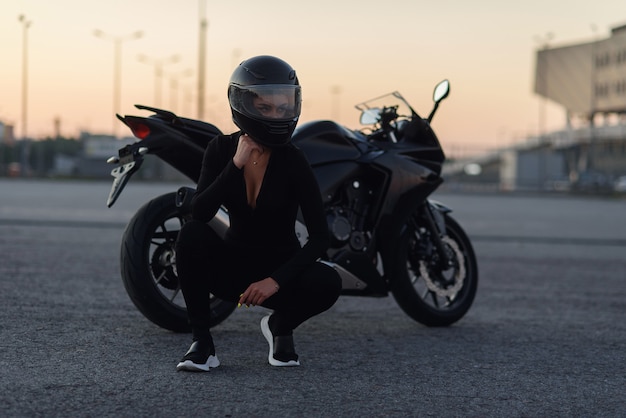 黒のタイトなボディスーツとフルフェイスヘルメットで美しい目を持つかなりバイカーの女性は、都市駐車場でスタイリッシュなスポーツバイクの近くに座っています。旅行とアクティブなライフスタイルのコンセプト。