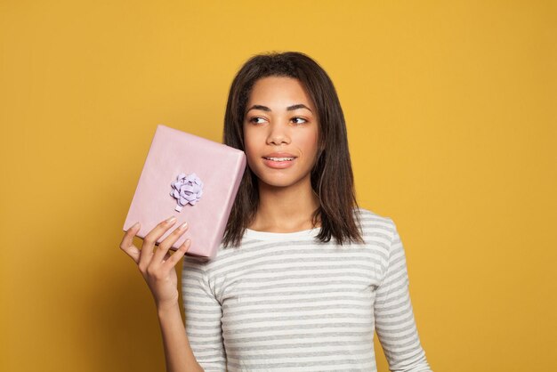 Красивая черная девушка с розовой коробкой подарков на красочном желтом фоне