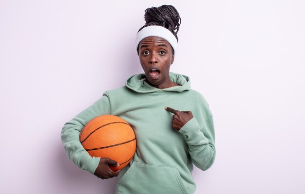 Pretty black woman looking shocked and surprised with mouth wide open, pointing to self. basketball concept