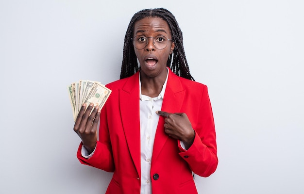 Pretty black woman looking shocked and surprised with mouth wide open, pointing to self. dollar banknotes concept