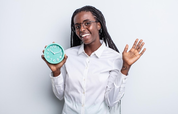 Pretty black woman smiling happily, waving hand, welcoming and greeting you. alarm clock concept