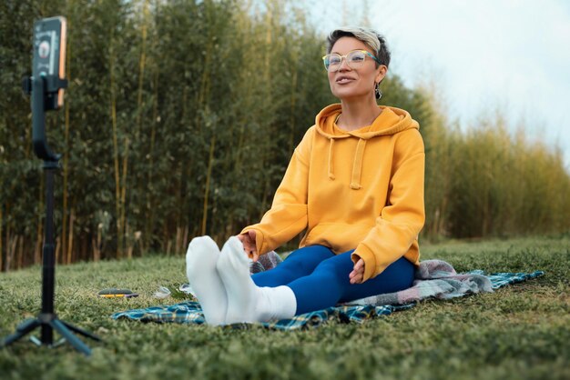 Photo pretty blogger girl in glasses and an orange hoodie is sitting in a park in nature streaming