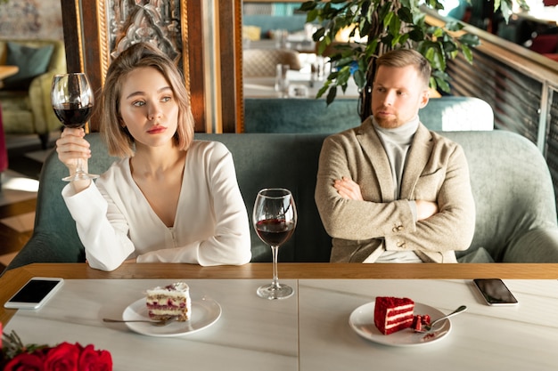 写真 近くに退屈な彼氏とレストランのテーブルのそばに座っている間、窓越しに見ている赤ワインのガラスとかなり金髪の女性