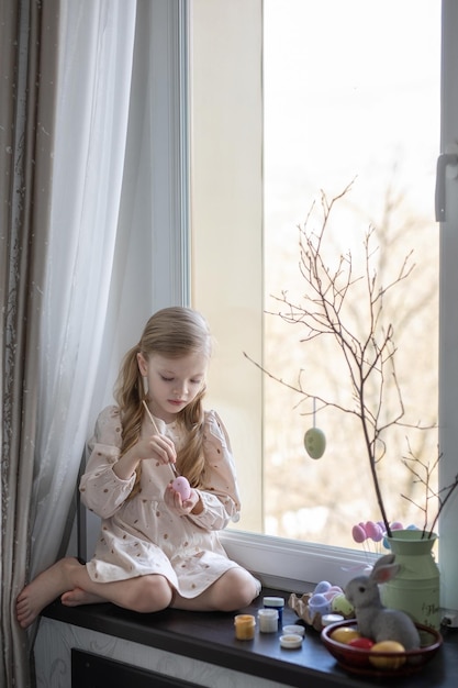 Pretty blond girl  is painting eggs for an Easter basket on the windowsill against the window