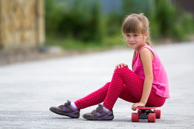 Pretty blond girl in pink clothing sitting on skateboard