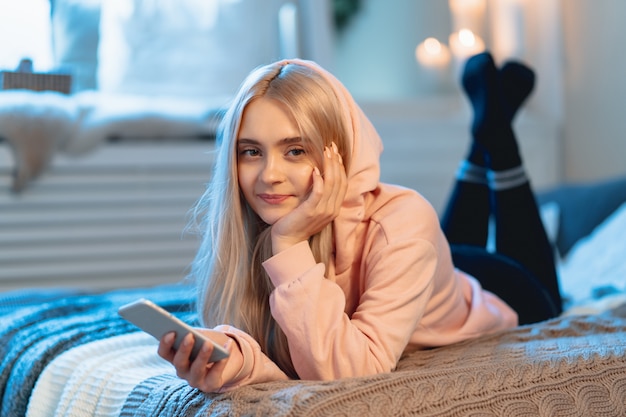 Довольно белокурая девушка в розовом с помощью смартфона лежит дома на кровати, пока общается со своими друзьями