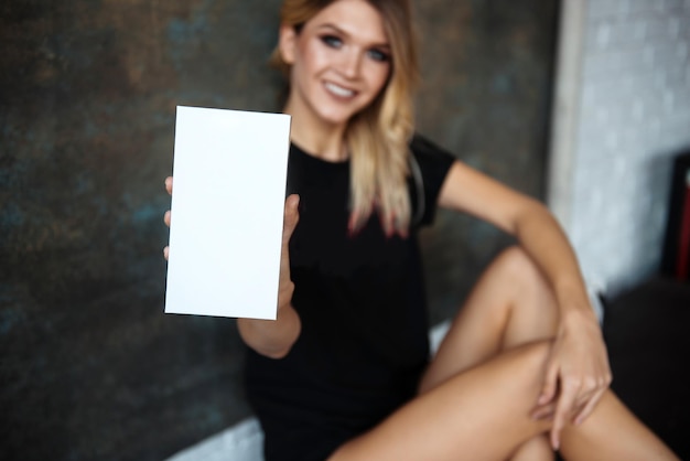 Pretty blond woman are blurred on a gray background and shows a blank white card in front of the camera Space for text mockup