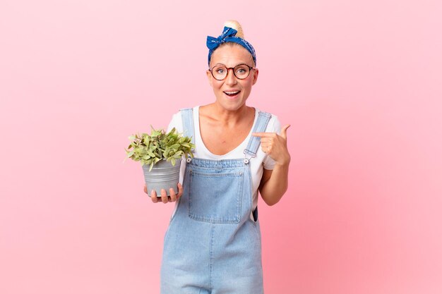 Pretty blond woman feeling happy and pointing to self with an excited and holding a plant