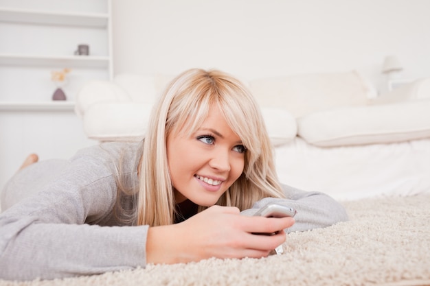 Pretty blond woman playing with cell phone lying down on a carpet