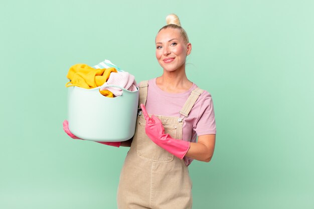 Pretty blond woman smiling cheerfully, feeling happy and pointing to the side washing clothes concept