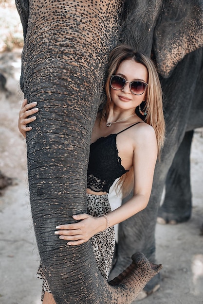 Pretty blond woman in stylish sunglasses and a dress with leopard print gently hugs elephant’s trunk; friendship concept.