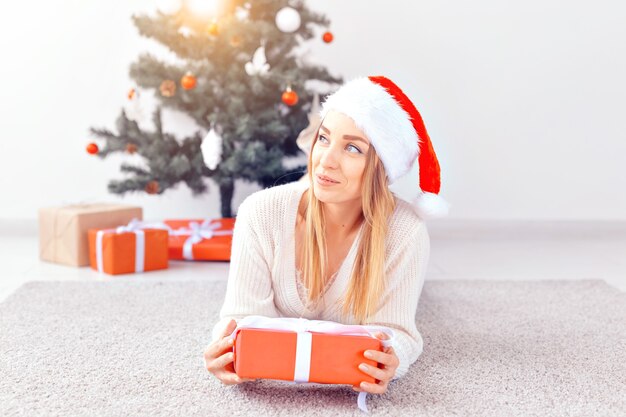 Pretty blond woman wearing a warm knitted sweater, lying on a carpet near decorated Christmas tree in a living room. A lot of presents under tree.