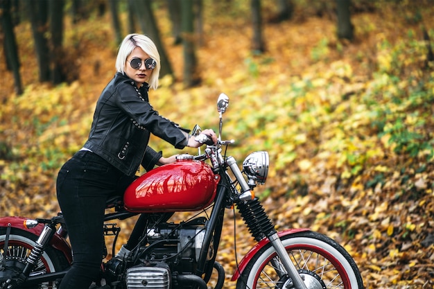 Pretty blonde biker girl sitting near red motorcycle on the road in the forest