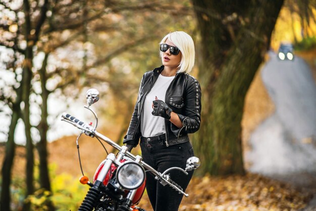 Pretty blonde biker girl in sunglasses with red motorcycle on the road in the forest